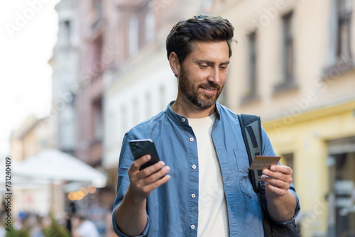 Mature bearded tourist with credit card and smartphone on evening city street