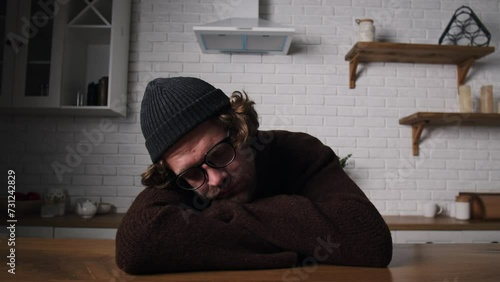 Tired young guy has no energy. Sleeping guy at dining room. Overworked man sleeping on folded hands in kitchen. Bored just fall asleep at home. Drunk slumber male in sweater and hat