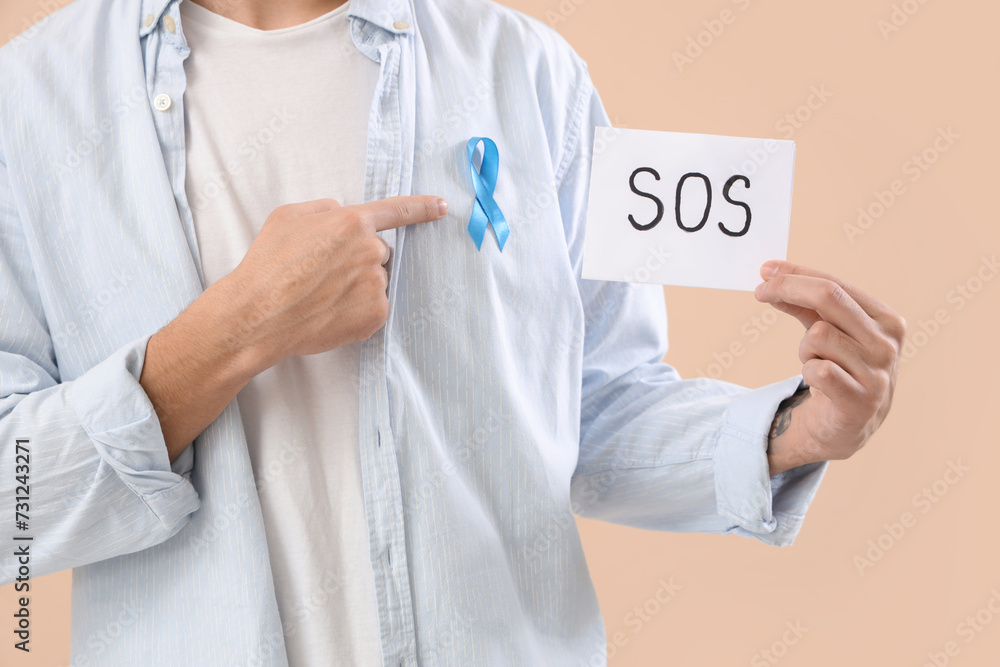 Young man holding paper with word SOS and blue ribbon on beige background, closeup. Prostate cancer awareness concept Young man holding paper with word SOS and blue ribbon on beige background, closeup. Prostate cancer awareness concept