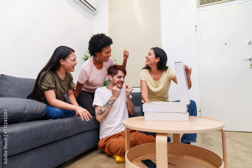 Joyful group of diverse young friends enjoying a pizza party at home. Excited man celebrates with cheerful women around a sofa. Joyful group of diverse young friends enjoying a pizza party at home. Excited man celebrates with cheerful women around a sofa.