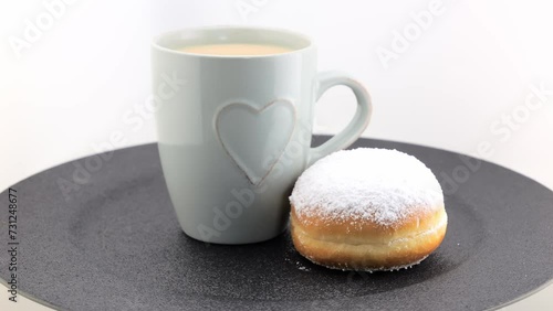 A cup of cappuccino and a freshly baked German doughnut with icing sugar, rotating on a black plate. Berlin donut (Berliner) or Krapfen. photo