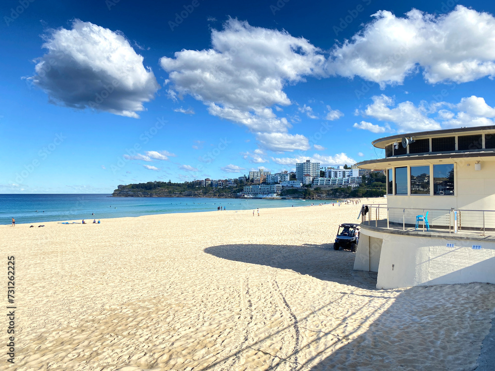 Obraz premium A day at the beach. Lifeguard hut on the sand to watch over people safety. Beach shore with sun and clouds. City’s buildings in the distance. 