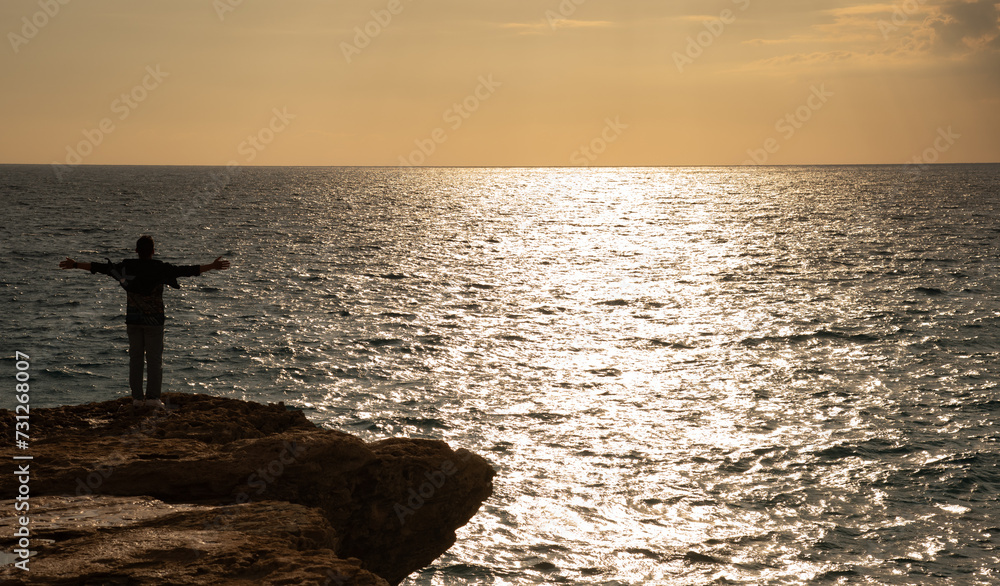 Silhouette of man raising hands or open arms at sunset at the edge of the cliff in the ocean