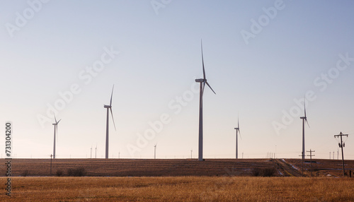 Wind turbines in a field in Kansas.  Side view. Utility lines and rural  road visible.