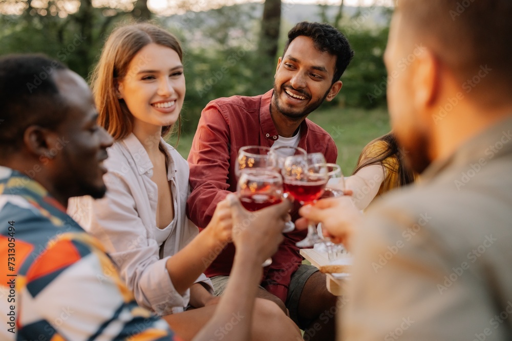 Multiracial group of happy friends sitting around table drinking wine talking at dinner party in evening summer forest Multiracial group of happy friends sitting around table drinking wine talking at dinner party in evening summer forest