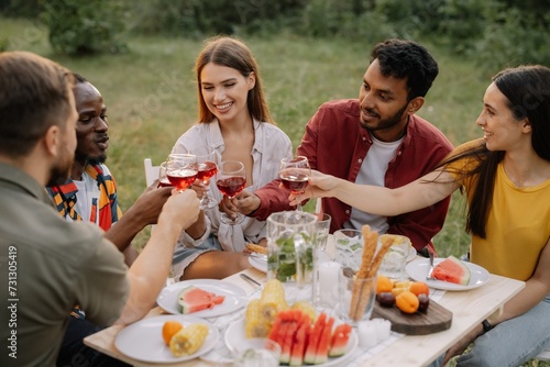 Multiracial group of happy friends sitting around table drinking wine talking at dinner party in evening summer forest