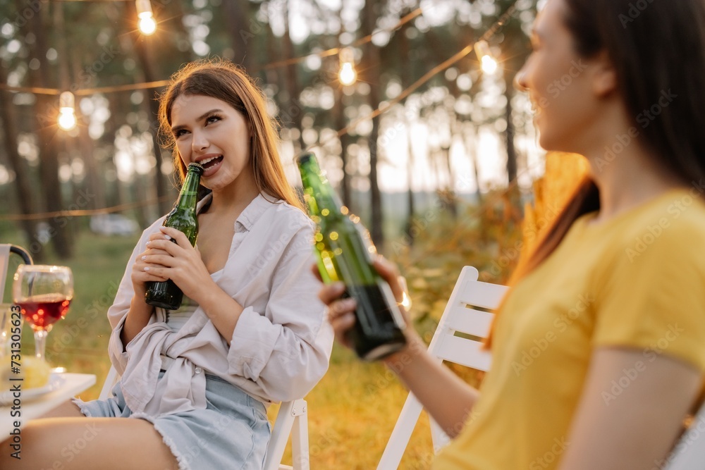 Funny hipster woman opens beer with teeth at party with friends in forest decorated with hanging lamps