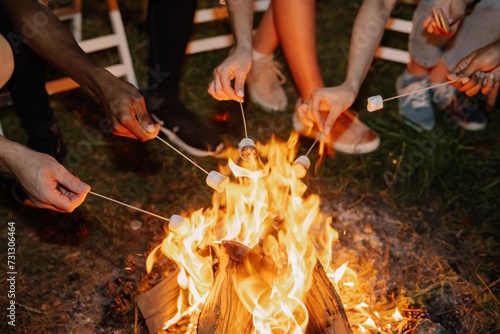 Group of multiracial restful friends roasting marshmallows while sitting by bonfire