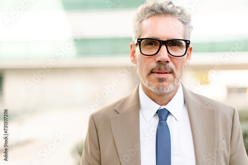 A senior businessman in glasses with grey hair poses confidently outdoors, his sharp beige suit and blue tie signaling a polished and authoritative presence. The concept of leadership and expertise