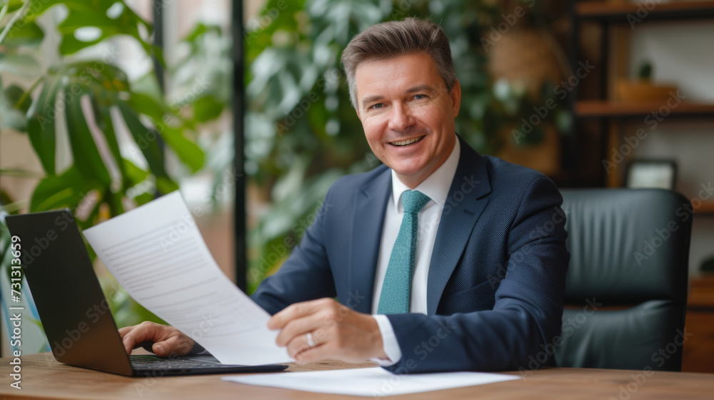 A smiling businessman in a suit is sitting at an office desk, holding a document with a laptop open in front of him. A smiling businessman in a suit is sitting at an office desk, holding a document with a laptop open in front of him.