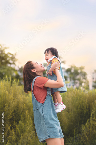 Happy Asian mother plays with daughter and holding girl in hands at park with nice sky, Baby smile and laughing,  Family concept.