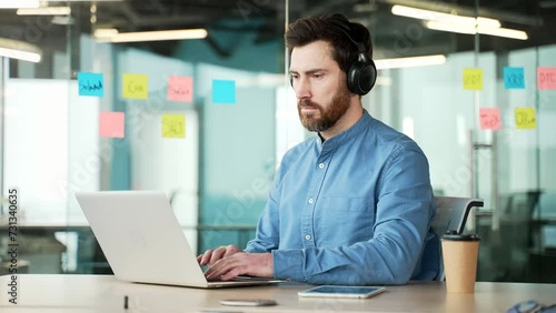 A focused IT specialist in wireless headphones works on a laptop sitting at a workplace in a business office. Serious bearded coder developer is programming, busy with a project on the computer photo