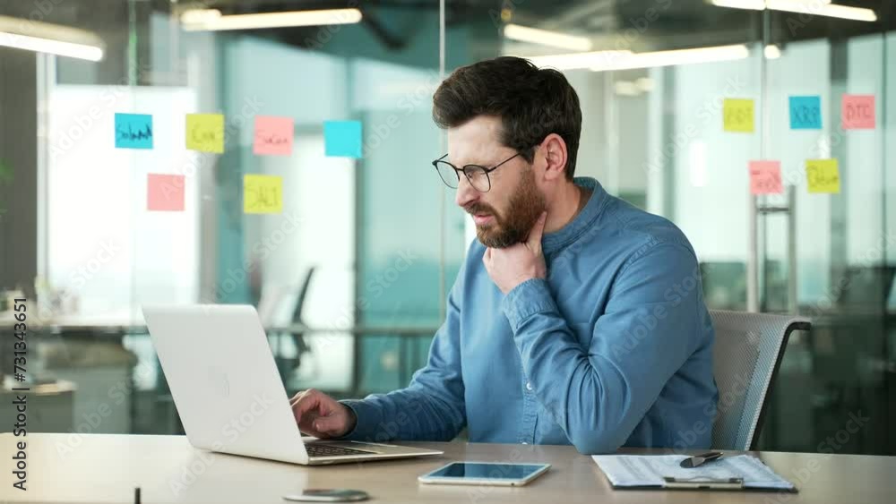 A sick employee suffering from a sore throat while working on a laptop while sitting at a workplace in a business office. A bearded man has a cold, virus or flu. Upset male massaging a sore spot A sick employee suffering from a sore throat while working on a laptop while sitting at a workplace in a business office. A bearded man has a cold, virus or flu. Upset male massaging a sore spot