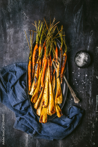 Roasted, glazed carrots and parsnips arranged on a dark oval platter with serving fork, and blue linen napkin on dark background photo