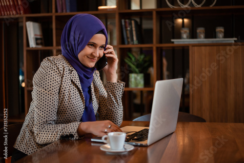 Successful smiling Arab woman in hijab working inside modern office, business woman using a smart phone Muslim woman using laptop at work