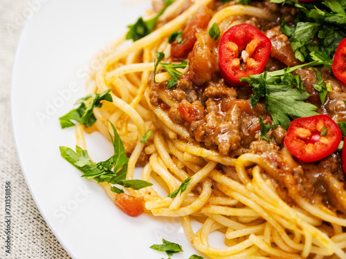 Spaghetti Bolognese dish with pasta, red sauce, minced beef, red peppers and green fresh herbs on white plate and light color table cloth. High quality products in a tasty Italian style dish.