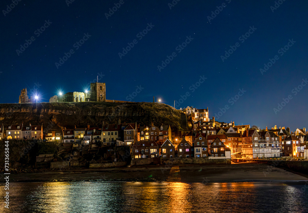 Whitby harbour at night. Abbey and church on the hill. Houses lights reflect on the water.