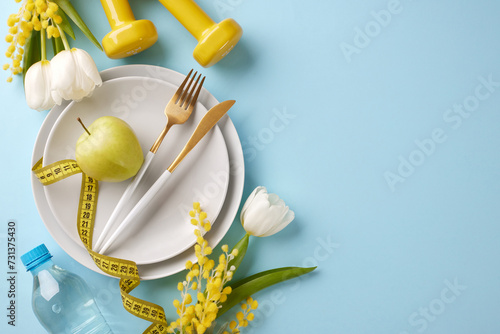 Slimming into spring: revitalize your body and mind. Top view shot of plates, cutlery, apple, yellow dumbbells, measure tape, water, fresh flowers on pastel blue background with advert space