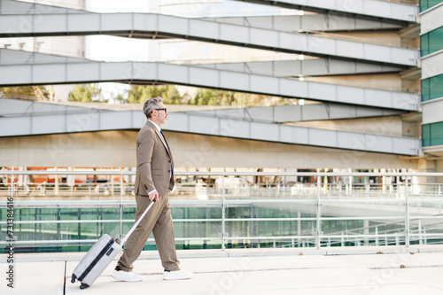A seasoned businessman is walking purposefully with a rolling suitcase, exemplifying the mobility and fast-paced nature of modern corporate life, side view with office on background
