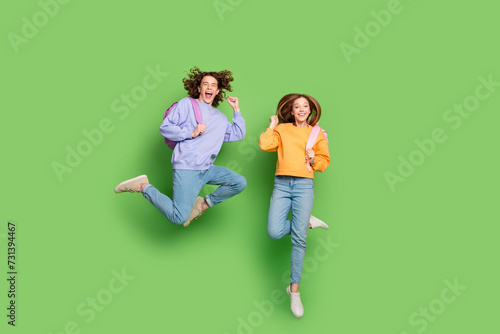 Full size photo of two excited cheerful people carry rucksack jump raise fists isolated on green color background