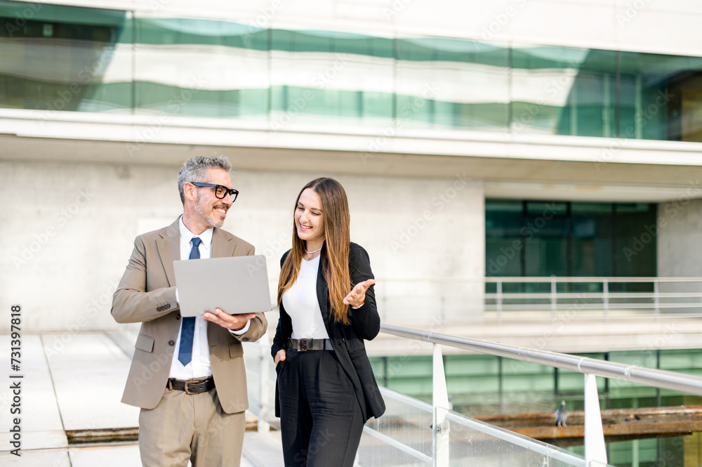 A senior businessman with grey hair and glasses shares a lighthearted moment with a younger female colleague while discussing work on a laptop outdoors, showcasing a mentorship or collaboration theme A senior businessman with grey hair and glasses shares a lighthearted moment with a younger female colleague while discussing work on a laptop outdoors, showcasing a mentorship or collaboration theme