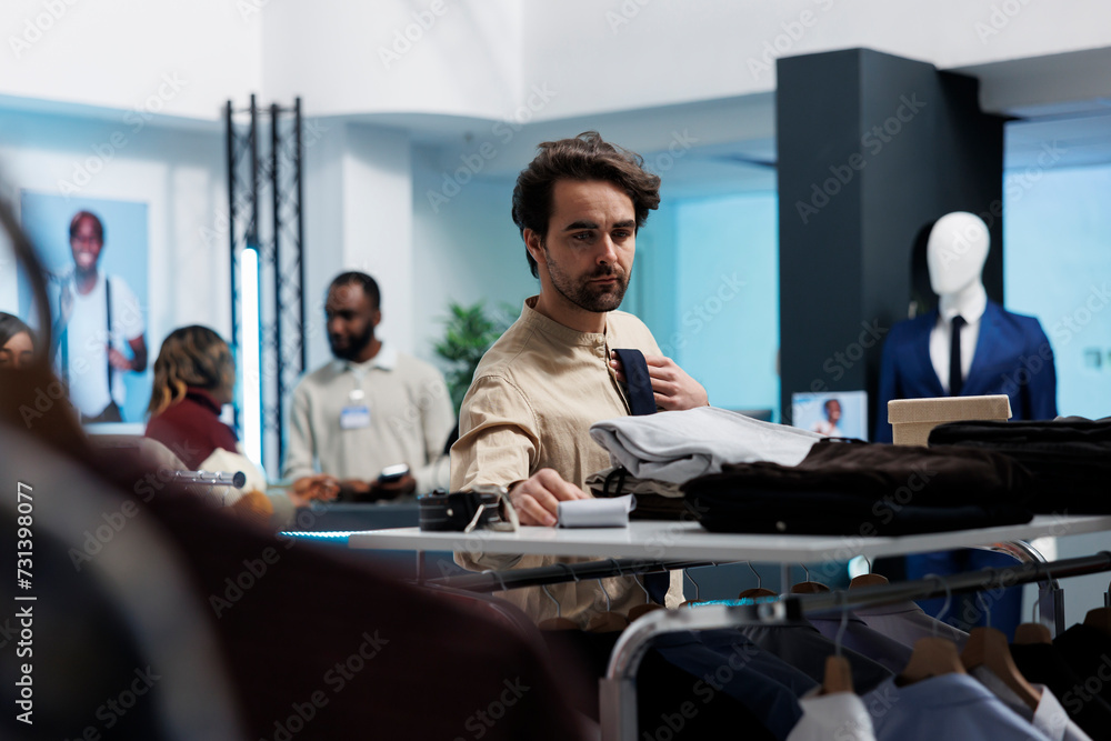 Clothing store client browsing accessory shelf while choosing tie in fashion boutique. Young caucasian man selecting formal outfit while shopping in mall menswear department