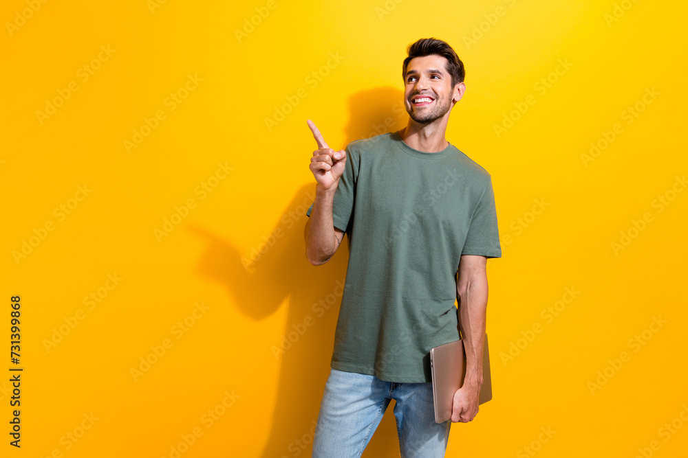 Portrait of good mood guy wear stylish t-shirt hold laptop directing look at promo empty space isolated on yellow color background Portrait of good mood guy wear stylish t-shirt hold laptop directing look at promo empty space isolated on yellow color background