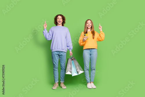 Full body photo of two funky people hold mall bags smart phone direct finger up empty space isolated on green color background © deagreez