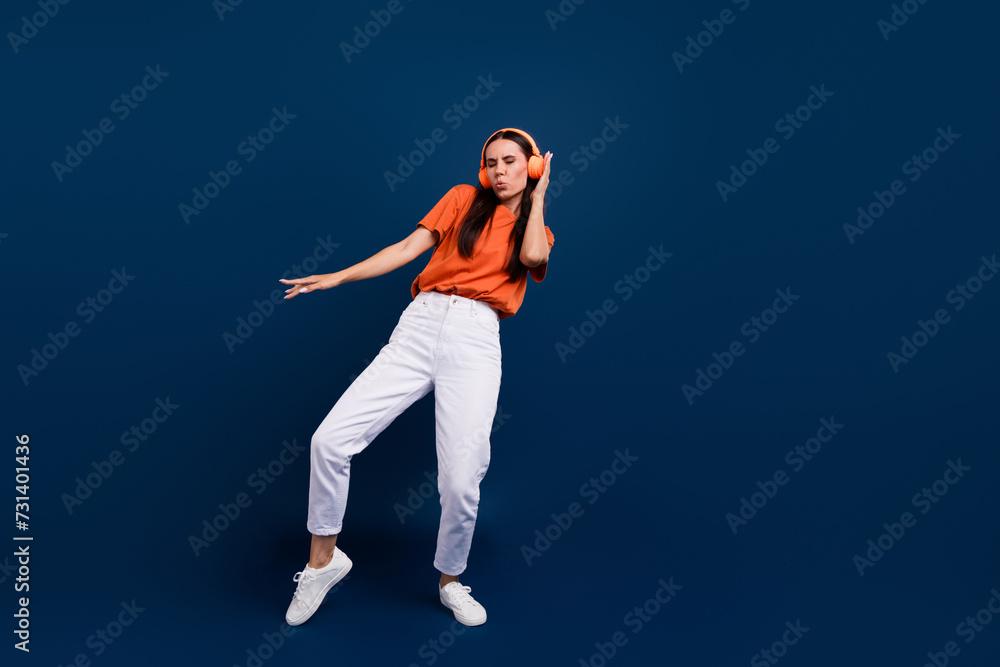 Full length photo of good mood excited woman dressed orange t-shirt earphones enjoying disco empty space isolated dark blue color background Full length photo of good mood excited woman dressed orange t-shirt earphones enjoying disco empty space isolated dark blue color background