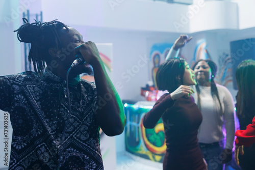 African american man singing in microphone while dancing with friends in crowded nightclub. Young clubber holding mic while clubbing partying at discotheque social gathering