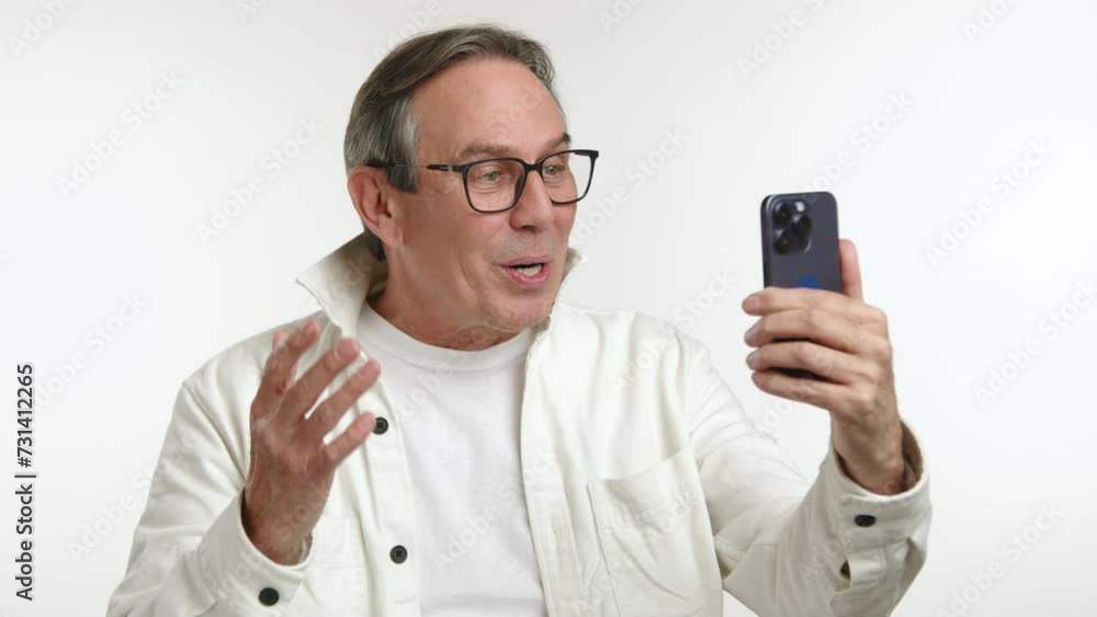 Cheerful mature man engaging in a video call, waving happily to the screen of his smartphone, dressed in a casual white jacket on a clean background. Camera 8K RAW. Cheerful mature man engaging in a video call, waving happily to the screen of his smartphone, dressed in a casual white jacket on a clean background. Camera 8K RAW.