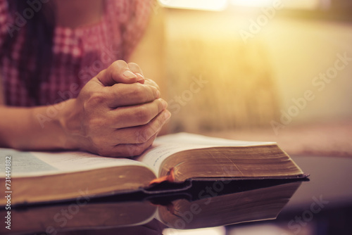 Close up of a woman hand  praying on  the open bible, blurred page on wooden table with window light and Bokeh, Christian devotional, spiritual or bible study concept background with copy space