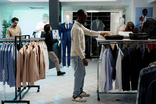 Stylish man searching for clothes and taking apparel from shelf in shopping mall department. African american shopper exploring apparel racks while choosing outfit in boutique