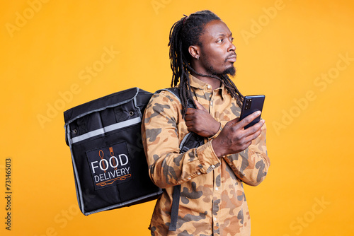 African american courier looking at customer address on smartphone takeout app before delivering order during lunch time. Deliveryman carrying thermal backpack full with takeaway food over photo