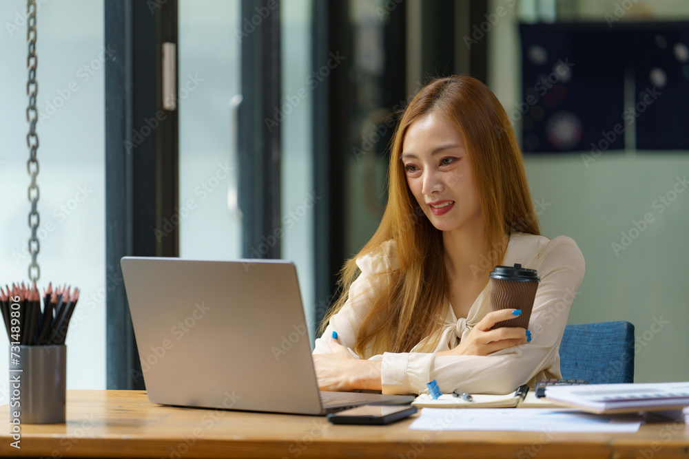 Successful Asian businesswoman smiling using a laptop computer and holding a coffee cup at the office. Confident Asia businesswoman sitting happily in the office. Successful Asian businesswoman smiling using a laptop computer and holding a coffee cup at the office. Confident Asia businesswoman sitting happily in the office.