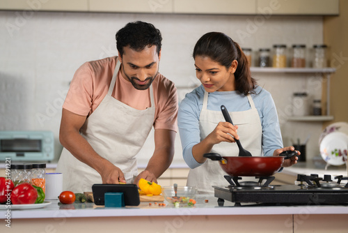 Indian couple at home preparing food by watching recipe on mobile phone app at kitchen during weekend - concept of relationship bonding, technology and teamwork