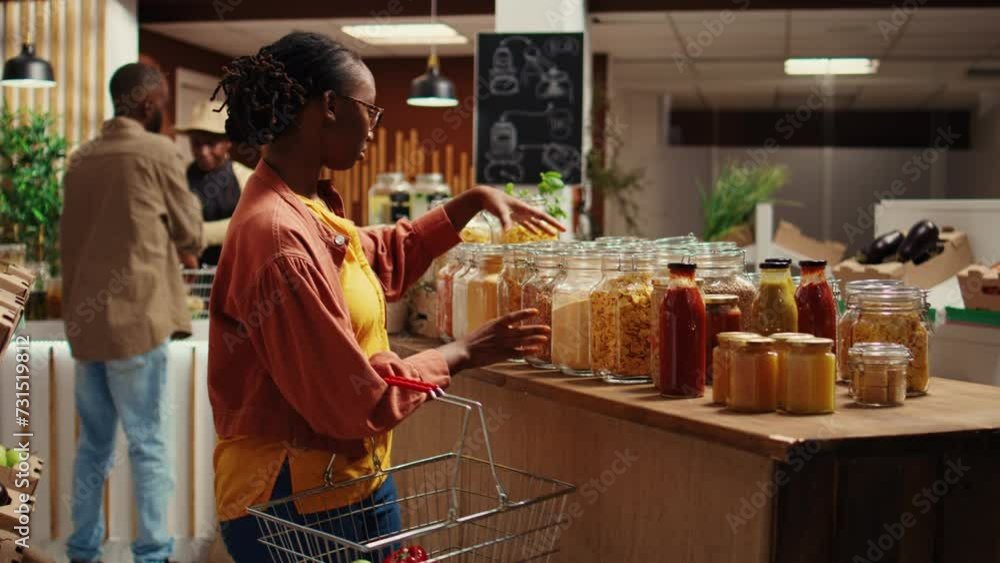 Female client checking variety of grains or pasta in reusable jars, looking to buy chemicals free pantry supplies from local farmers market. African american buyer shopping for bio items. Camera B. Female client checking variety of grains or pasta in reusable jars, looking to buy chemicals free pantry supplies from local farmers market. African american buyer shopping for bio items. Camera B.