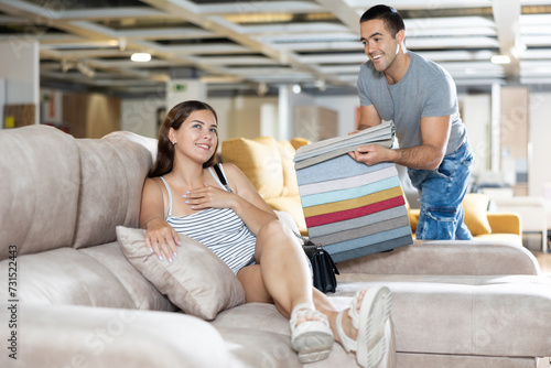 In sales area of furniture store,young woman is sitting on sofa with ottoman and testing it before buying.Sales consultant shows her set of upholstery fabrics for color selection