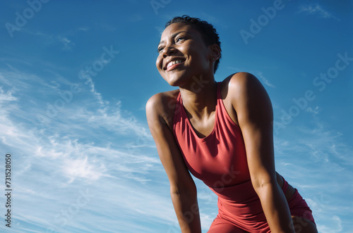 In the image, a woman is wearing a red tank top and is smiling while looking upwards. She has short hair and is crouching. The sky in the background is blue with some clouds