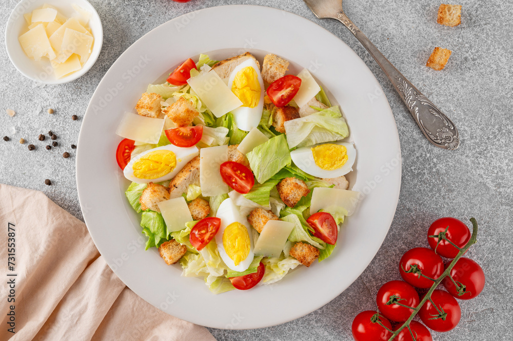 Caesar salad with lettuce, grilled chicken breast, boiled eggs, tomatoes, parmesan cheese and croutons in a ceramic plate on a concrete background. Top view. Caesar salad with lettuce, grilled chicken breast, boiled eggs, tomatoes, parmesan cheese and croutons in a ceramic plate on a concrete background. Top view.