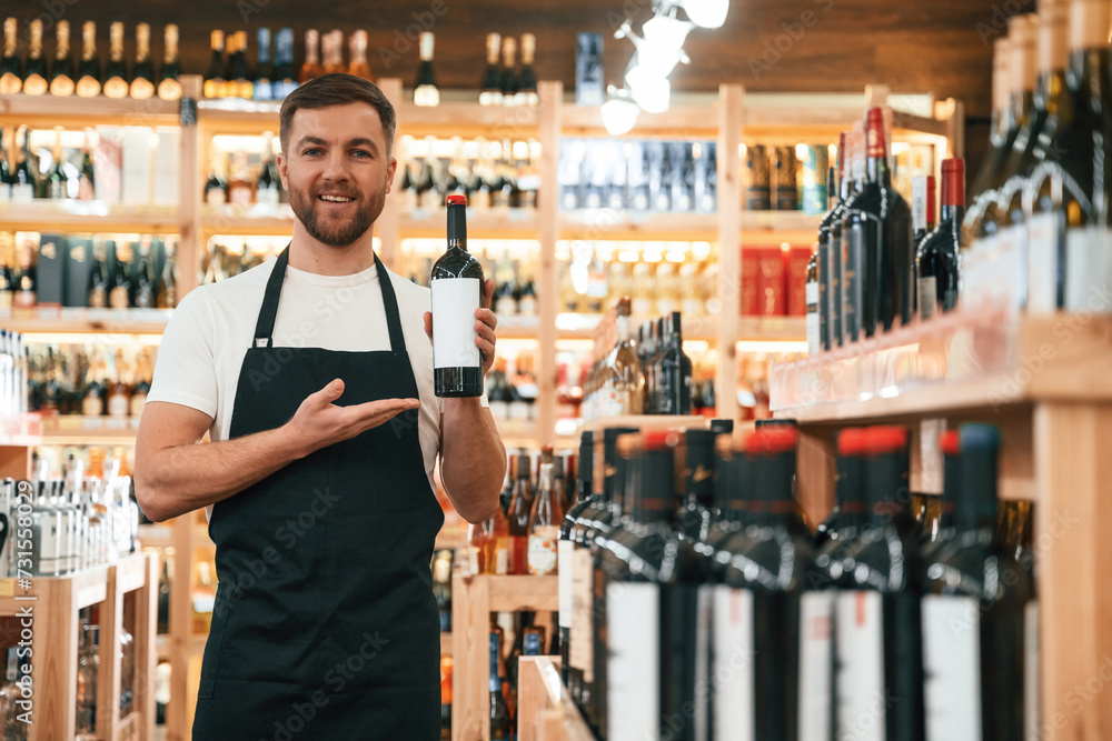 Man is holding bottle and smiling. Wine shop owner in white shirt and black apron