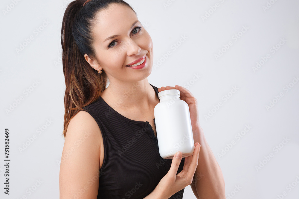 A cheerful caucasian woman, wearing a black sleeveless top, holding a white supplement bottle, against a light background, exuding a friendly vibe. Mockup for design. A cheerful caucasian woman, wearing a black sleeveless top, holding a white supplement bottle, against a light background, exuding a friendly vibe. Mockup for design.