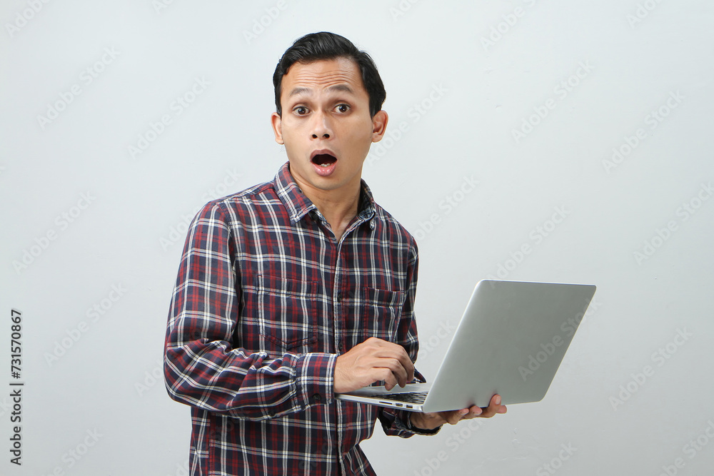 Shocked and wow expression of asian indonesian man holding laptop computer on isolated background Shocked and wow expression of asian indonesian man holding laptop computer on isolated background