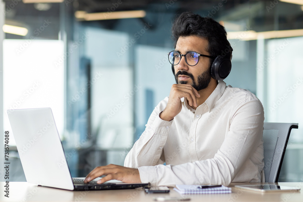 A man wearing headphones is sitting at a desk using a laptop computer A man wearing headphones is sitting at a desk using a laptop computer