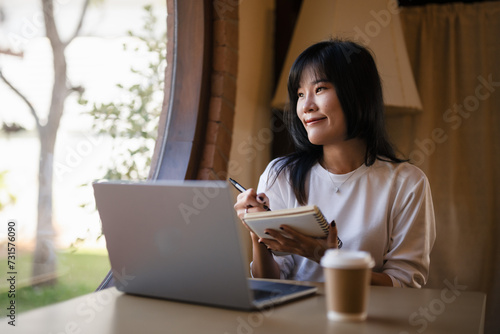 Cheerful young woman with a laptop and notebook, enjoying her work from home setup in a sunny room..