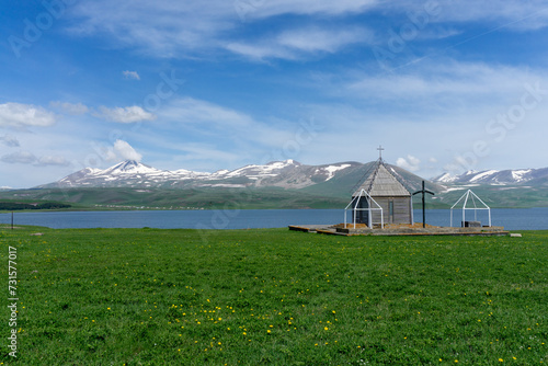  Small chapel near Paravani lake. Natural lake, blue water, clouds, snowy mountains and blue sky.