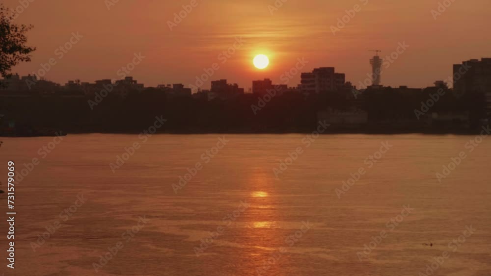A beautiful shot of the sunset setting against the city buildings in the background and the calm flowing river water in the foreground with a reflection of the vibrant orange sky. A beautiful shot of the sunset setting against the city buildings in the background and the calm flowing river water in the foreground with a reflection of the vibrant orange sky.