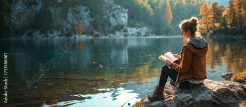 femme en train de lire, assise sur un rocher à côté d'un lac photo