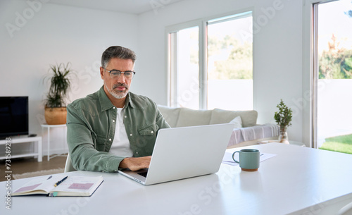 Serious older mature middle aged man wearing glasses looking at computer technology sitting at home table, using laptop hybrid working online, elearning, browsing web, searching online in living room. photo
