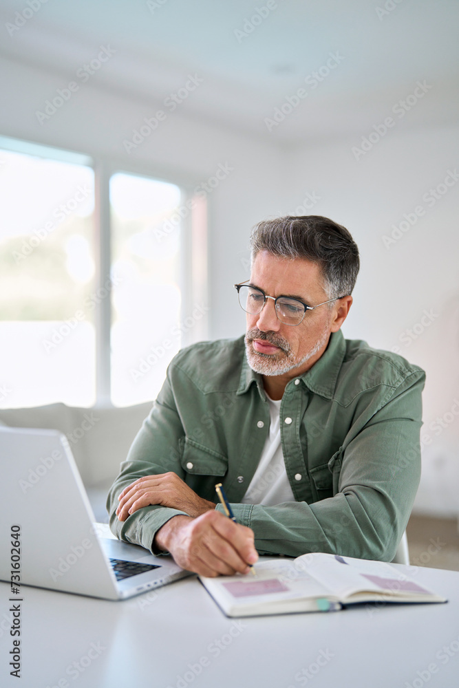 Serious older mature middle aged man wearing eyeglasses looking at computer technology sitting at table, using laptop hybrid working online, elearning, watching webinar writing notes. Vertical Serious older mature middle aged man wearing eyeglasses looking at computer technology sitting at table, using laptop hybrid working online, elearning, watching webinar writing notes. Vertical